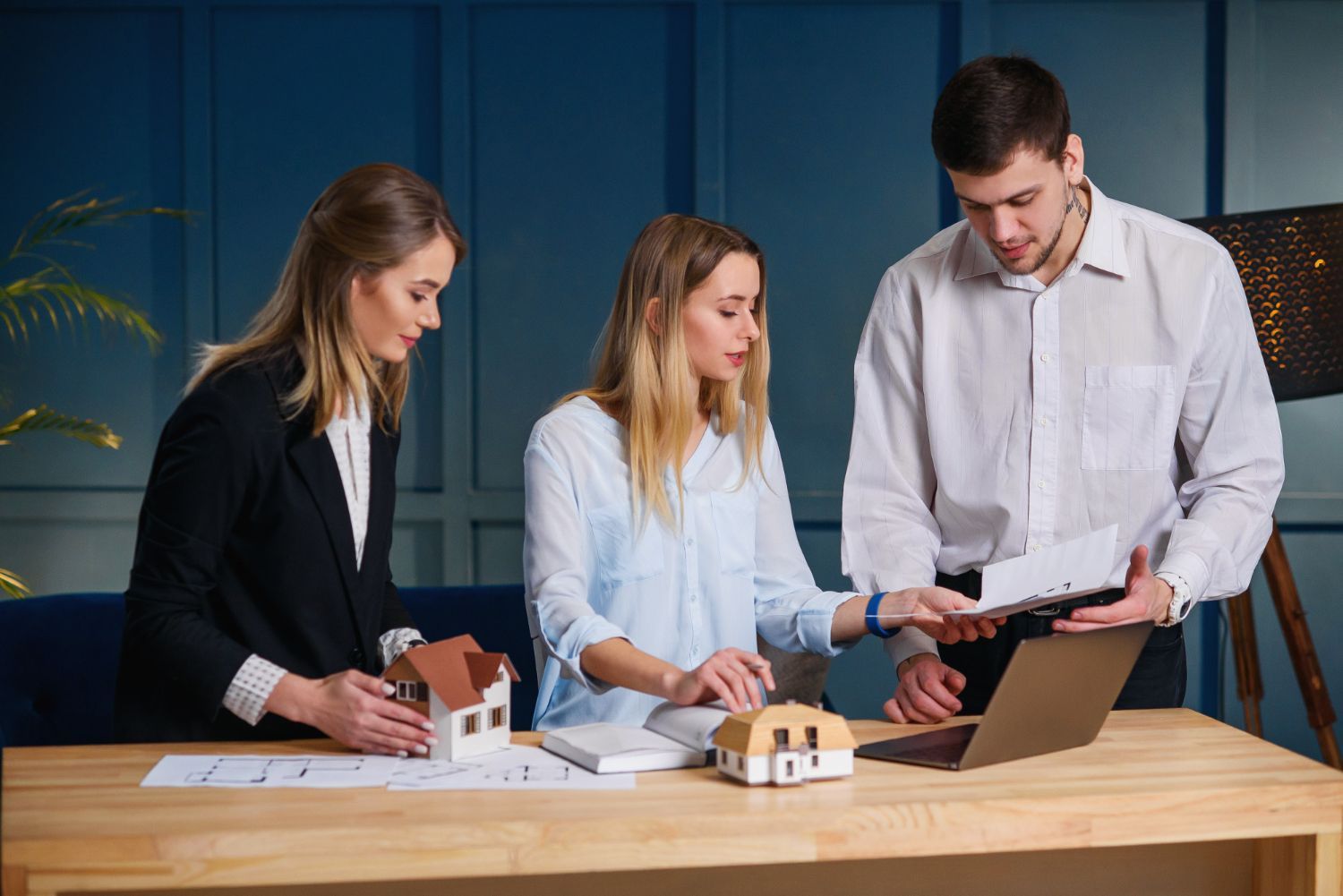 Three people review documents at a desk with model houses and a laptop.