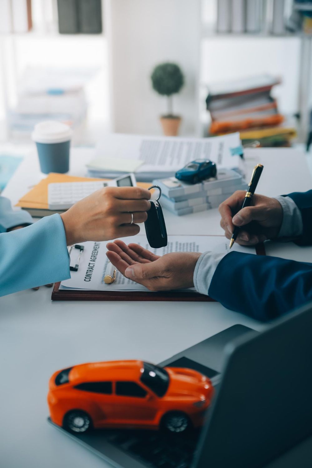 Person handing car keys to another person signing a contract. Orange toy car in the foreground.