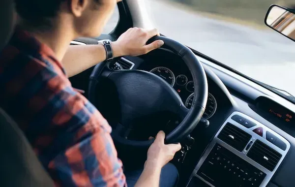 Person driving a car, hands on the steering wheel, wearing a plaid shirt.