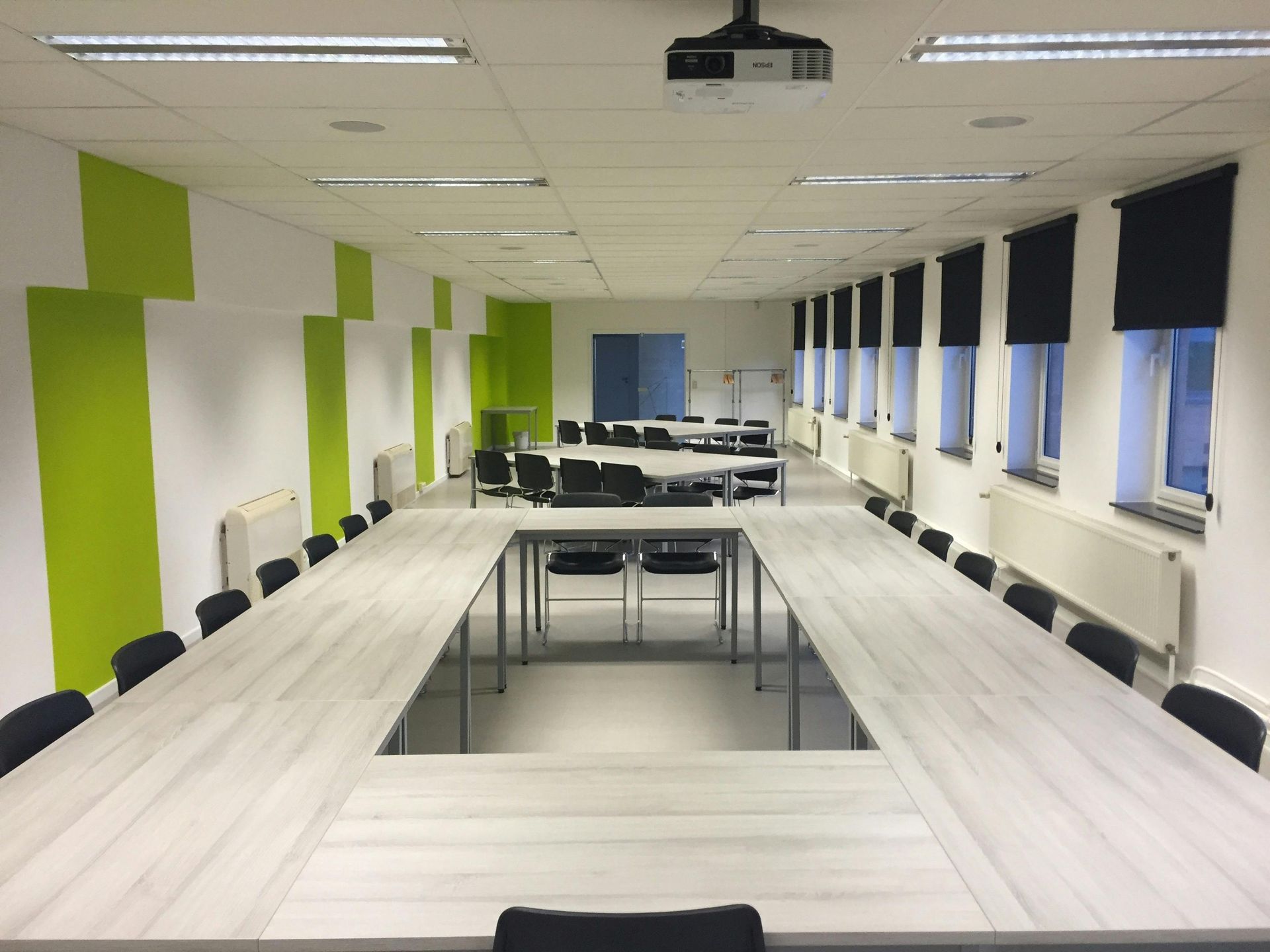 Conference room with long table, black chairs, white and green walls, and windows with black shades.