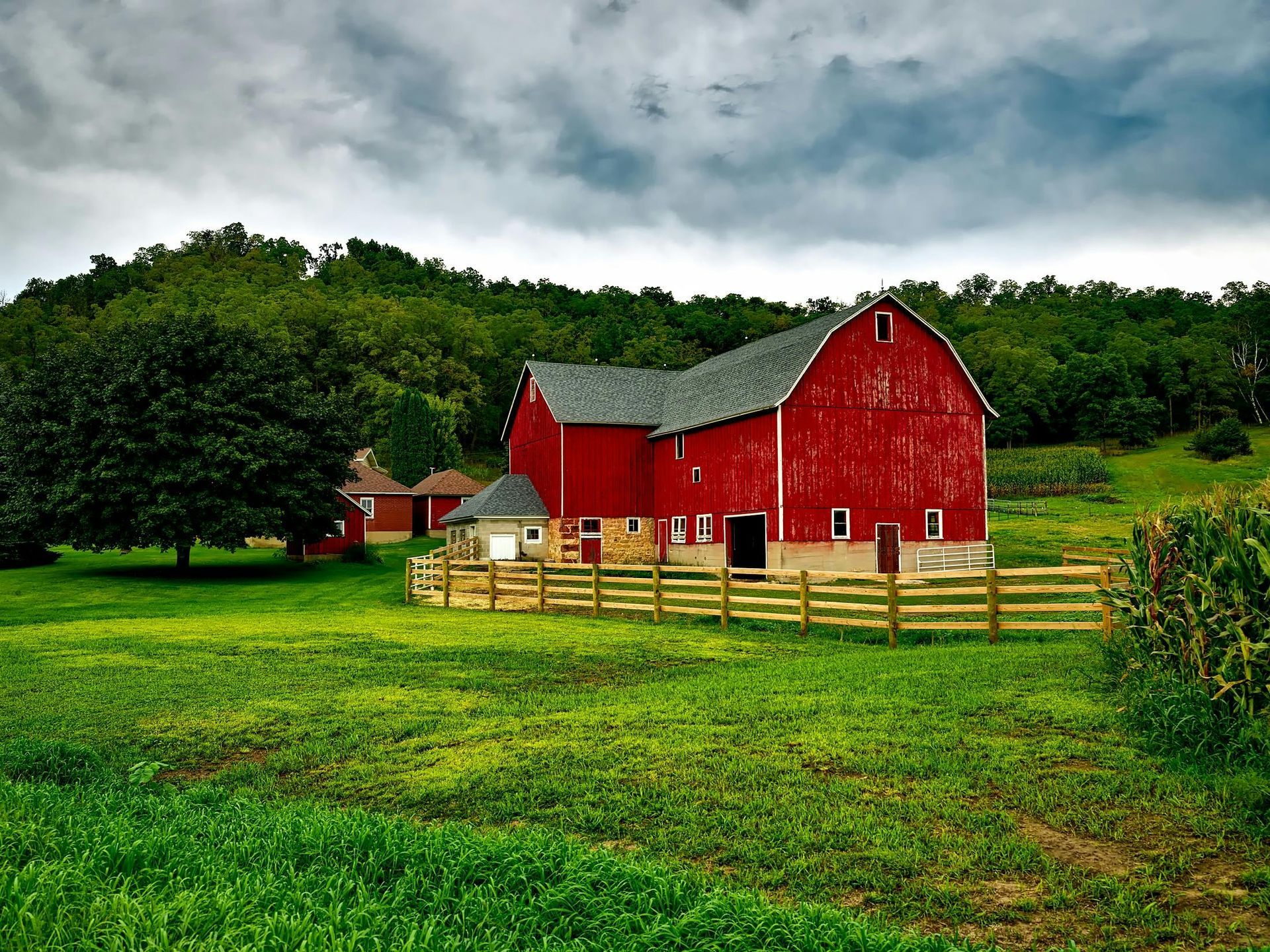 Red barn in a green field, wooden fence, trees in the background, cloudy sky.