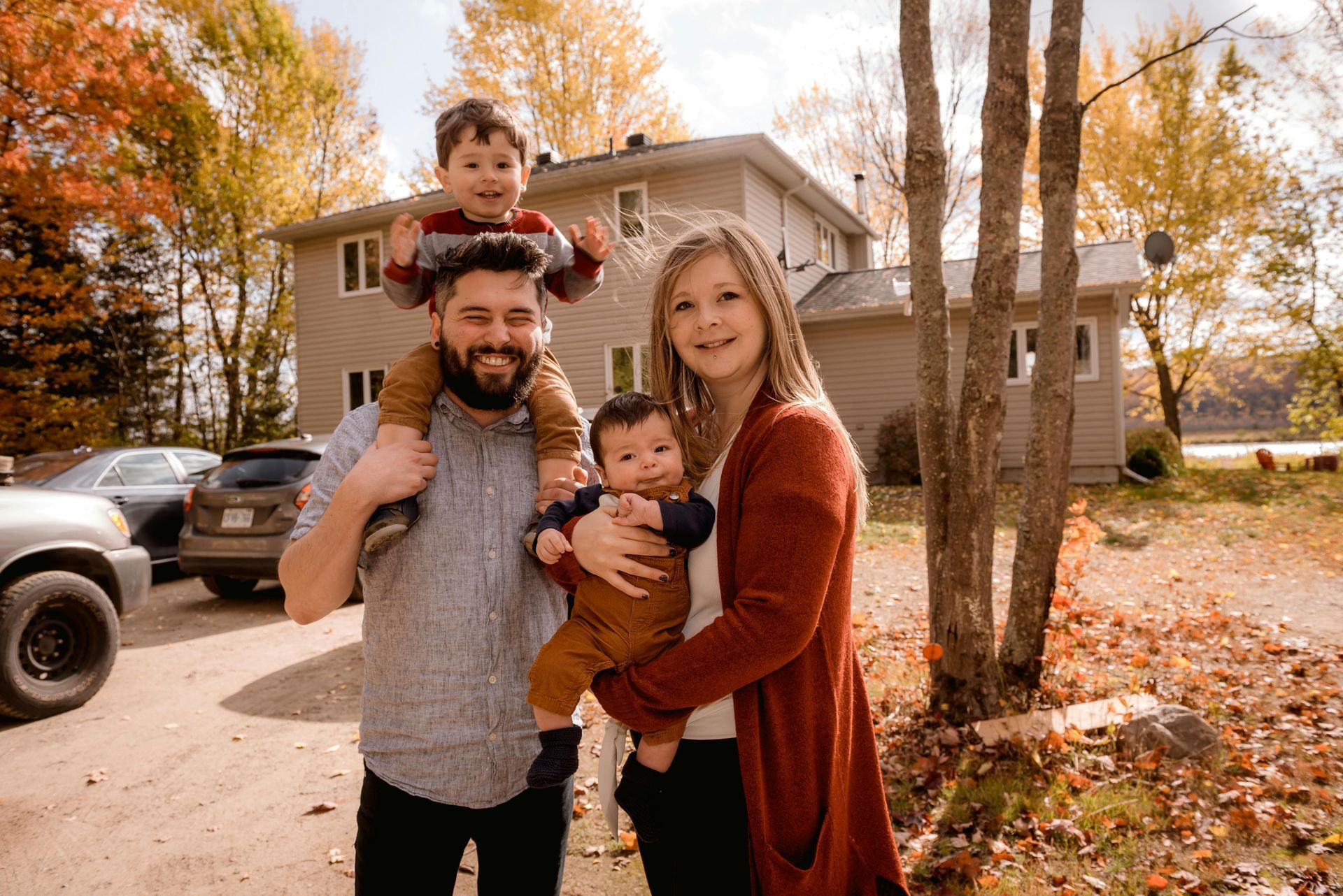 Family poses in front of a house: father with child on shoulders, mother holding a baby, autumn leaves.