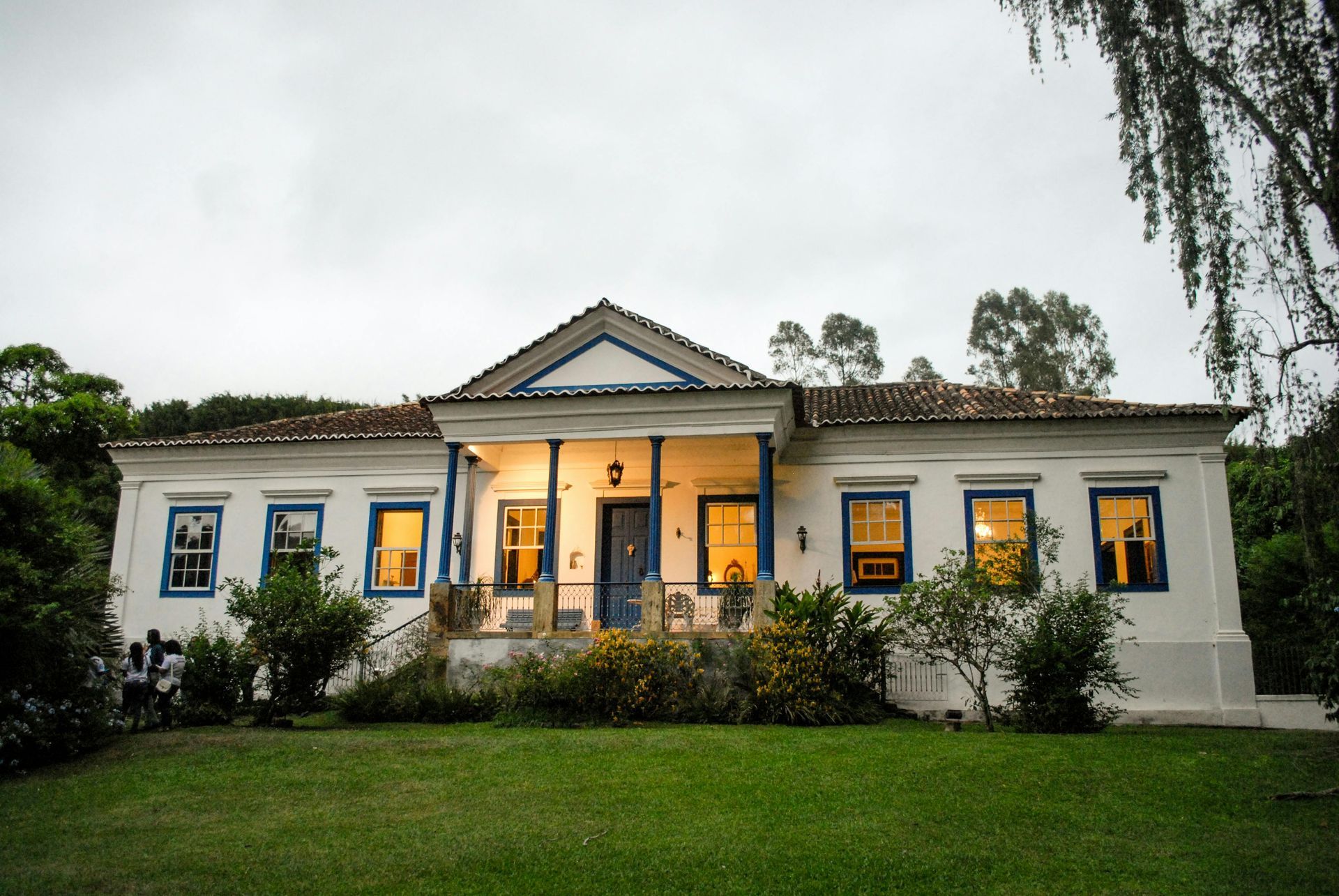 White house with blue trim, lights on in windows, green lawn, overcast sky.