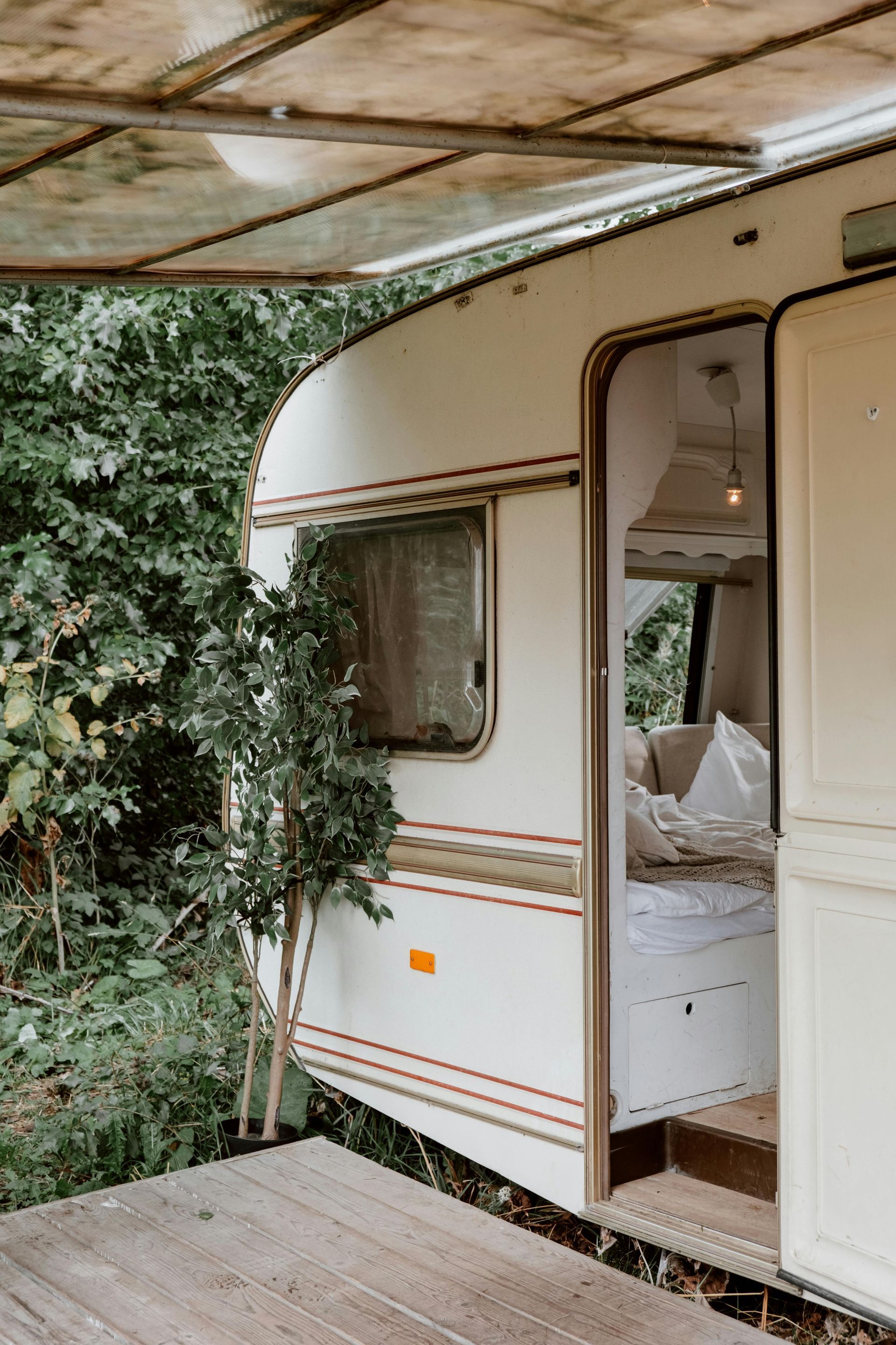 White campervan with open door, bed visible. Wooden deck, surrounded by greenery.