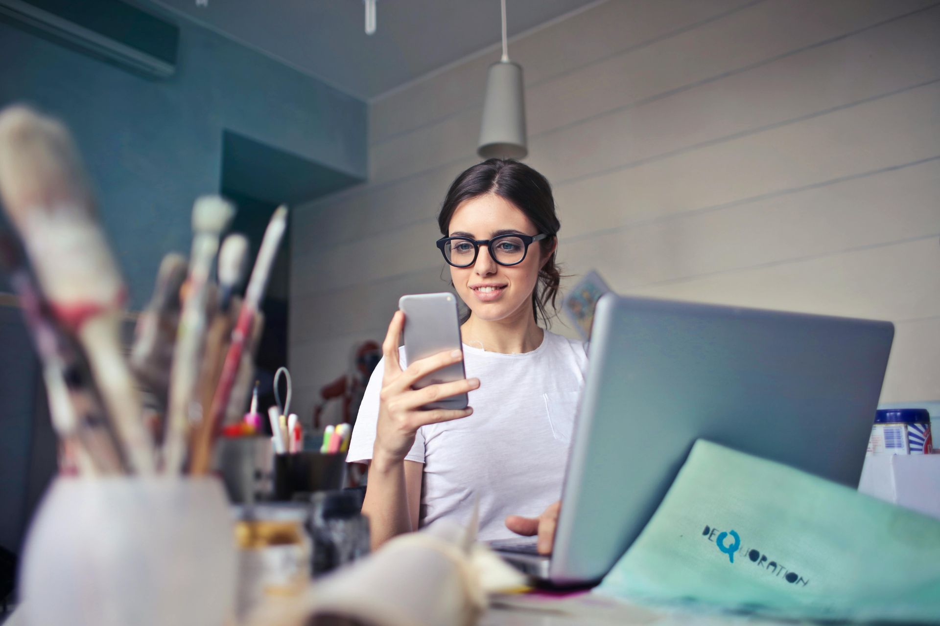 Woman wearing glasses, looking at phone, laptop open, art supplies in background.