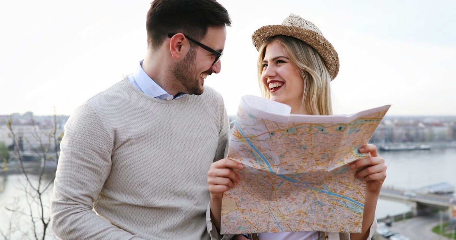 Couple looking at a map, smiling, outdoors; city and river in the background.