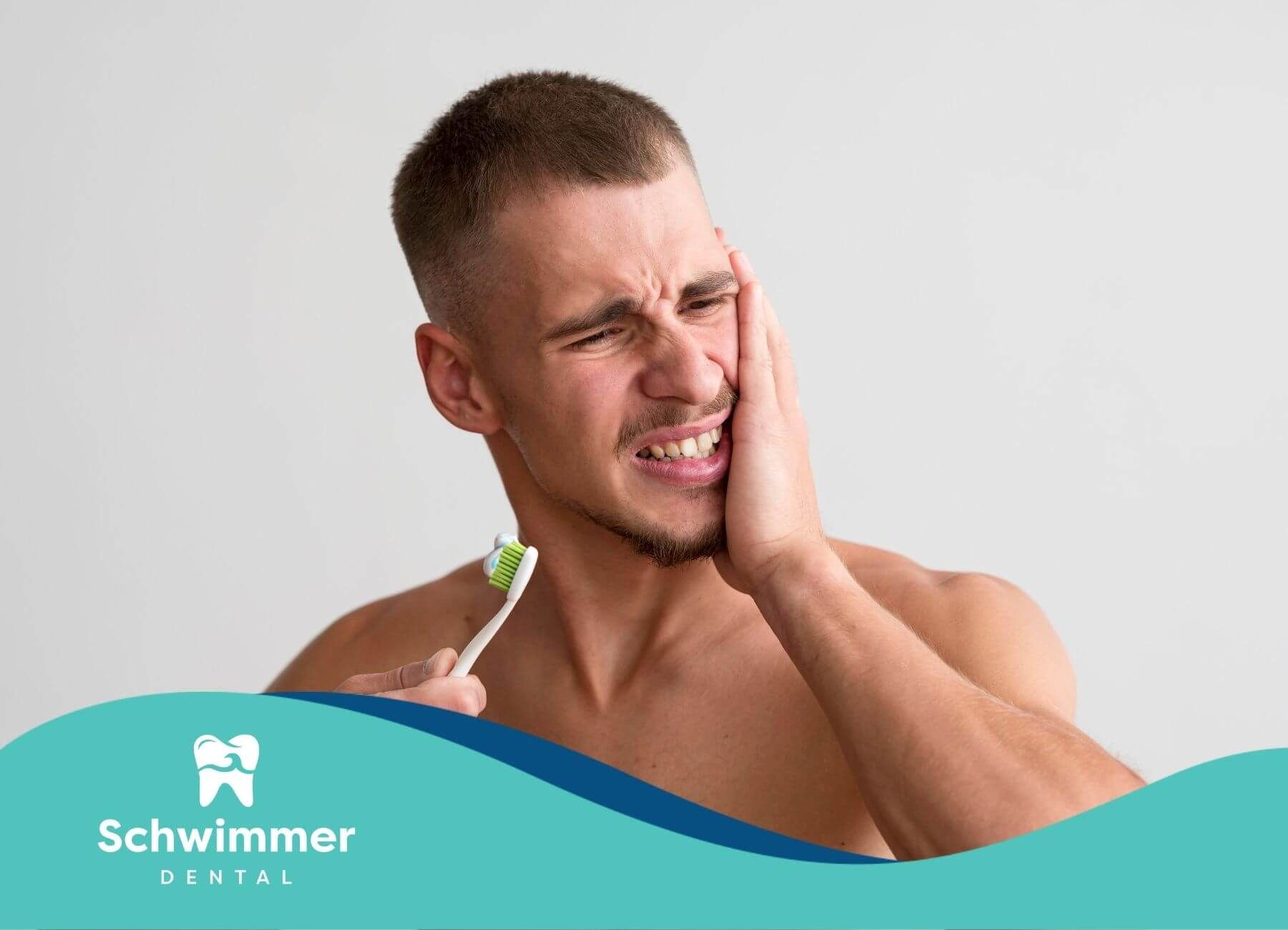 A man grimaces in pain while brushing his teeth, holding his jaw in discomfort due to a toothache.