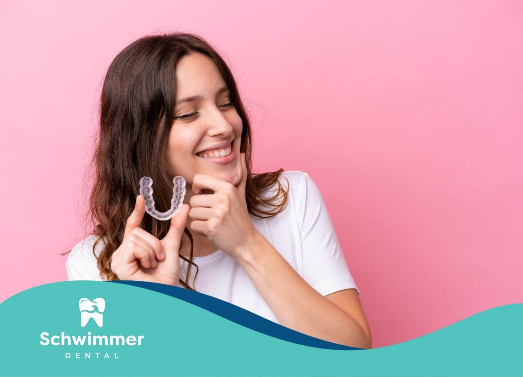Smiling woman holding a clear orthodontic retainer in front of pink background.