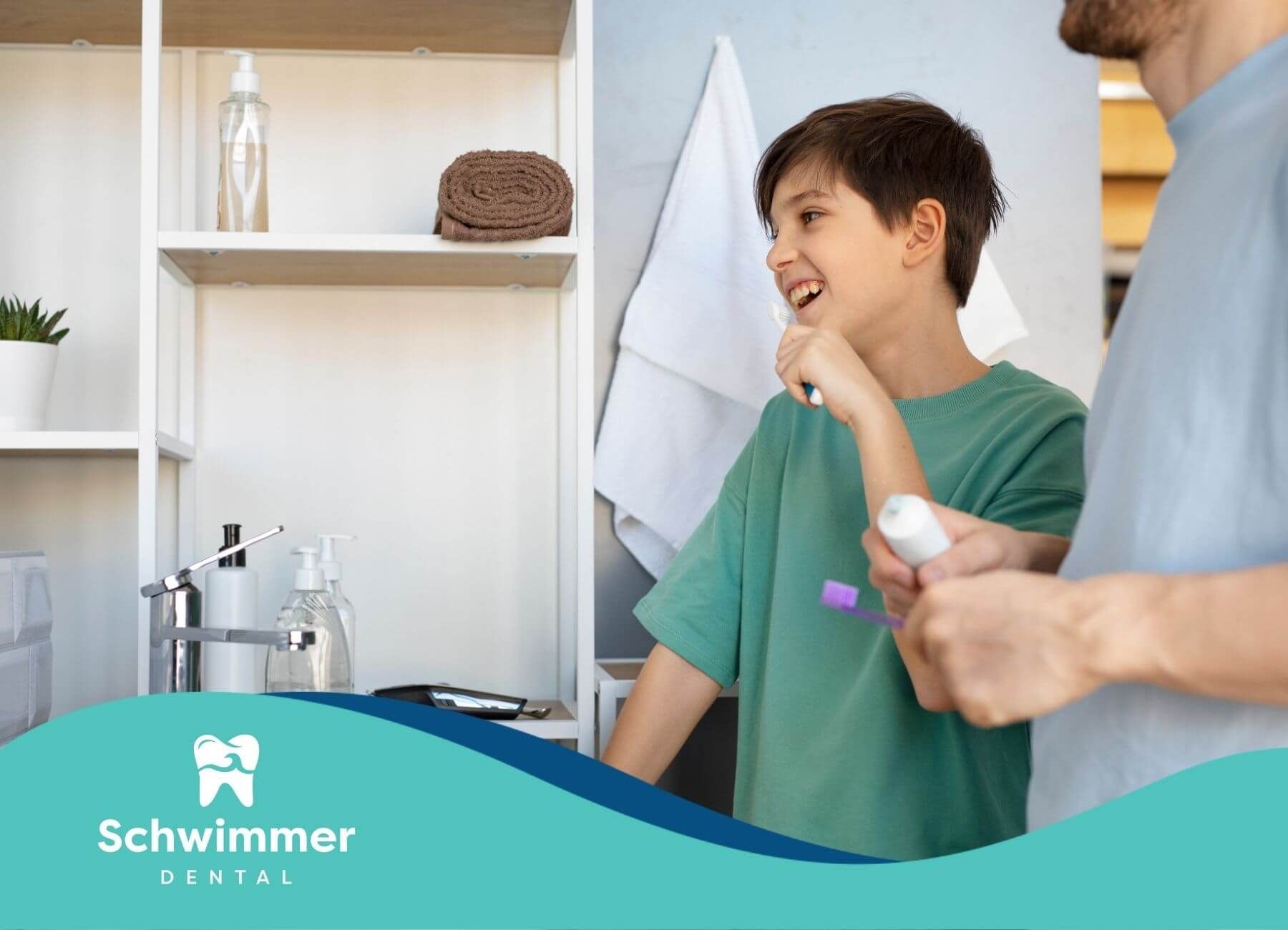 A boy brushes his teeth in a bathroom with his father, both smiling while holding their toothbrushes