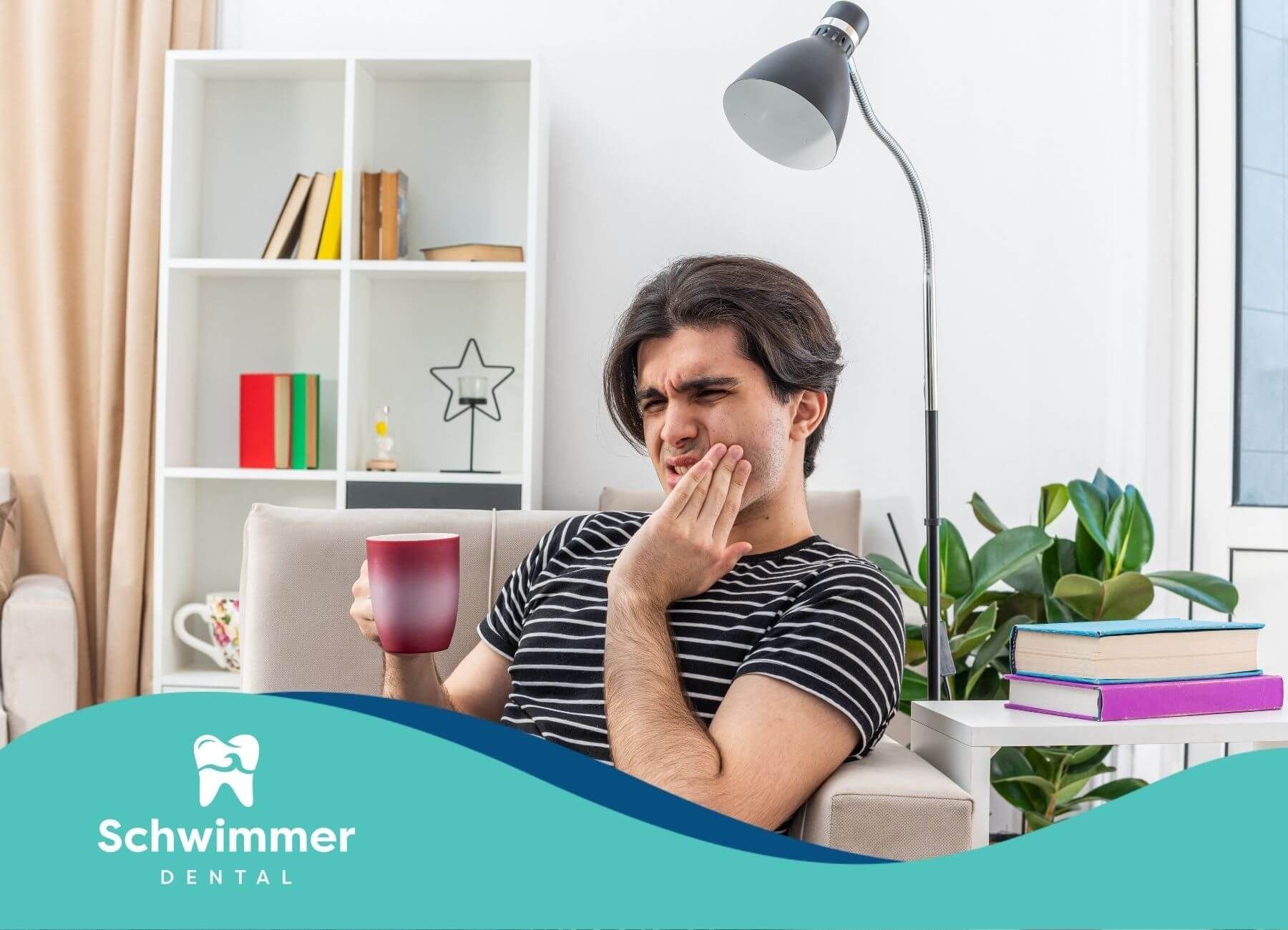 A man sitting on a couch, holding a cup, with a pained expression as they touch their cheek