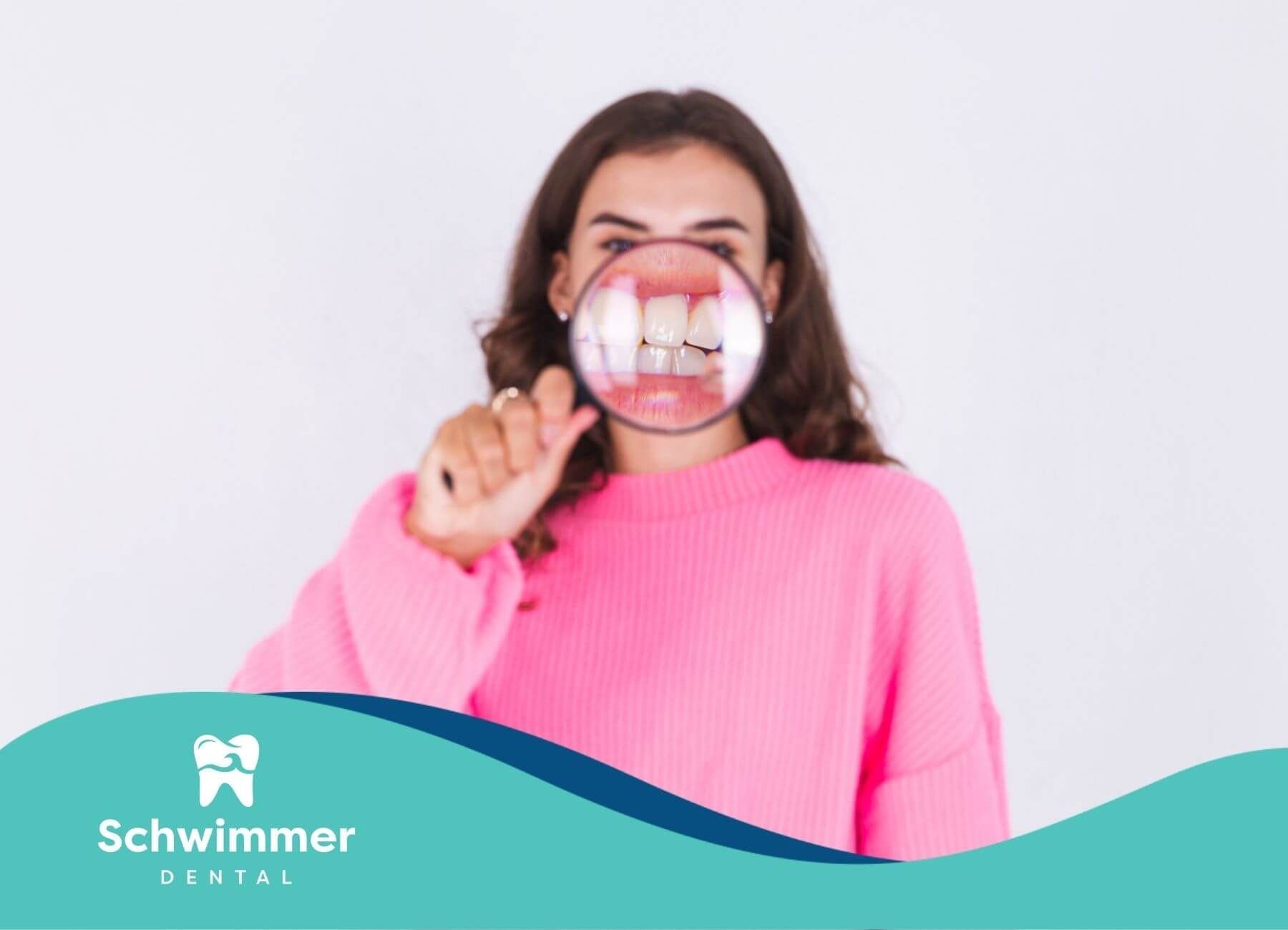 A woman in a pink sweater holding a magnifying glass over her mouth, inspecting her teeth.
