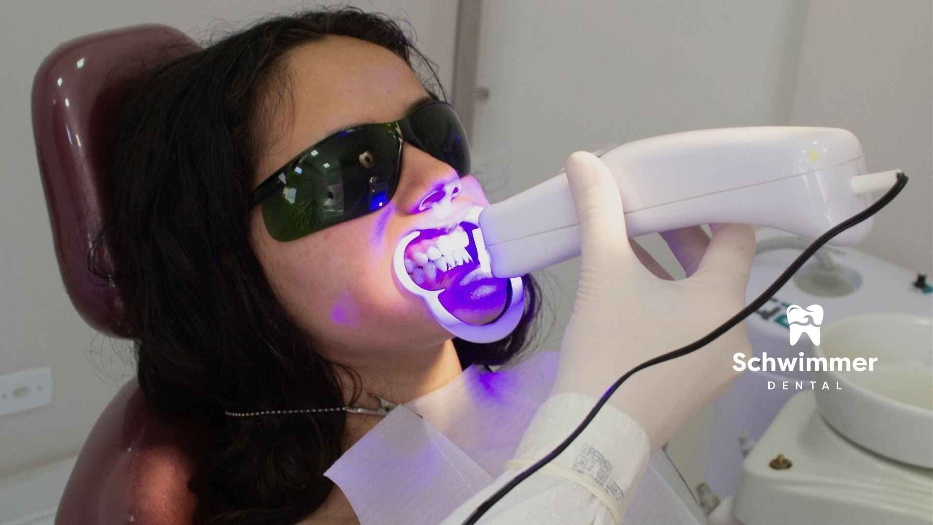 Woman at dentist, mouth open, receiving treatment with light instrument, wearing protective glasses.