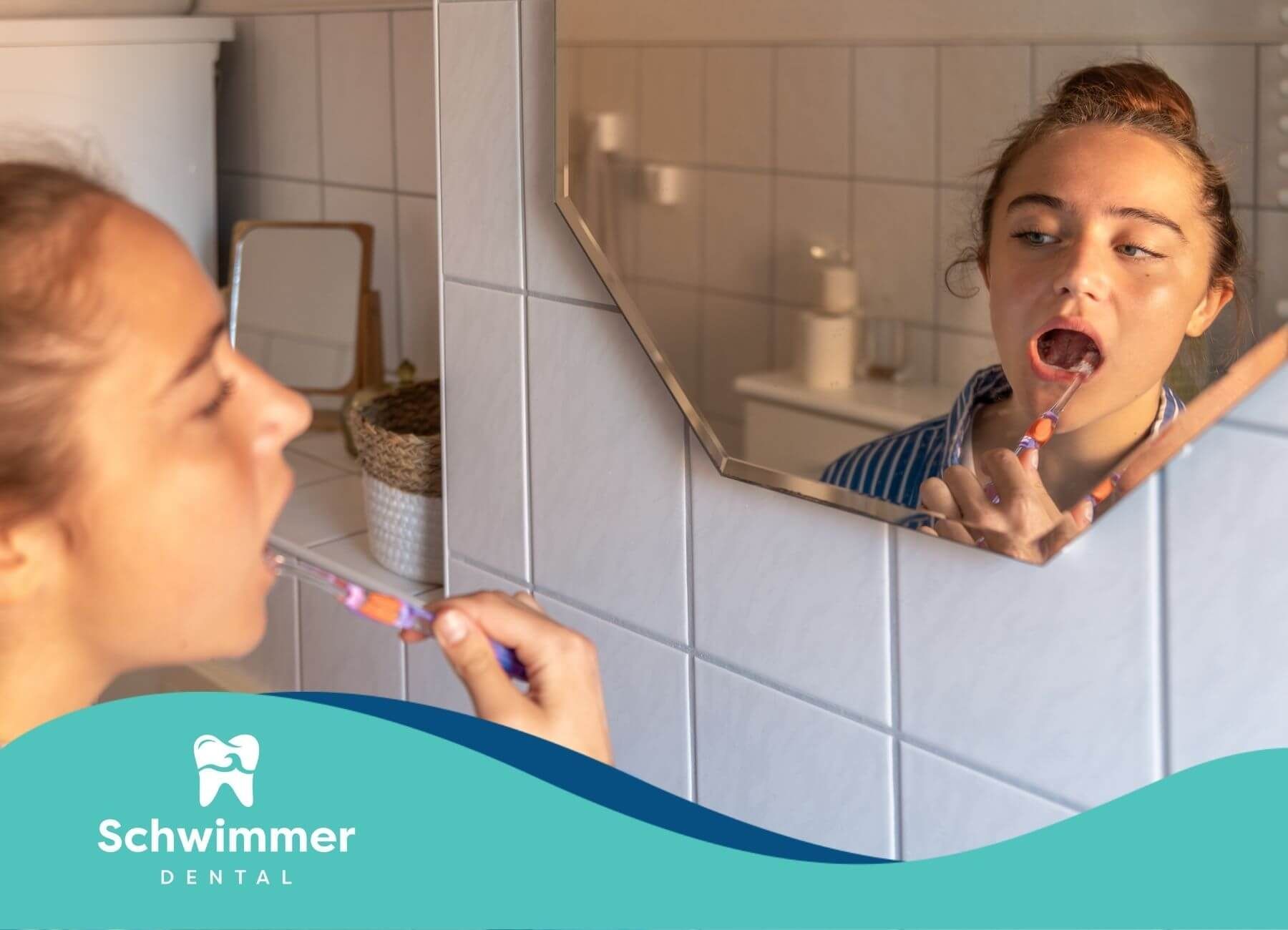 A woman brushes her teeth while looking at her reflection in the bathroom mirror, wearing pajamas.