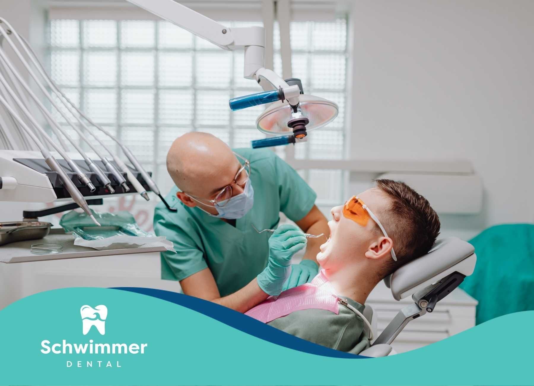 Dentist examining a patient's mouth in a dental office. Both wear protective gear; the patient is wearing orange glasses.
