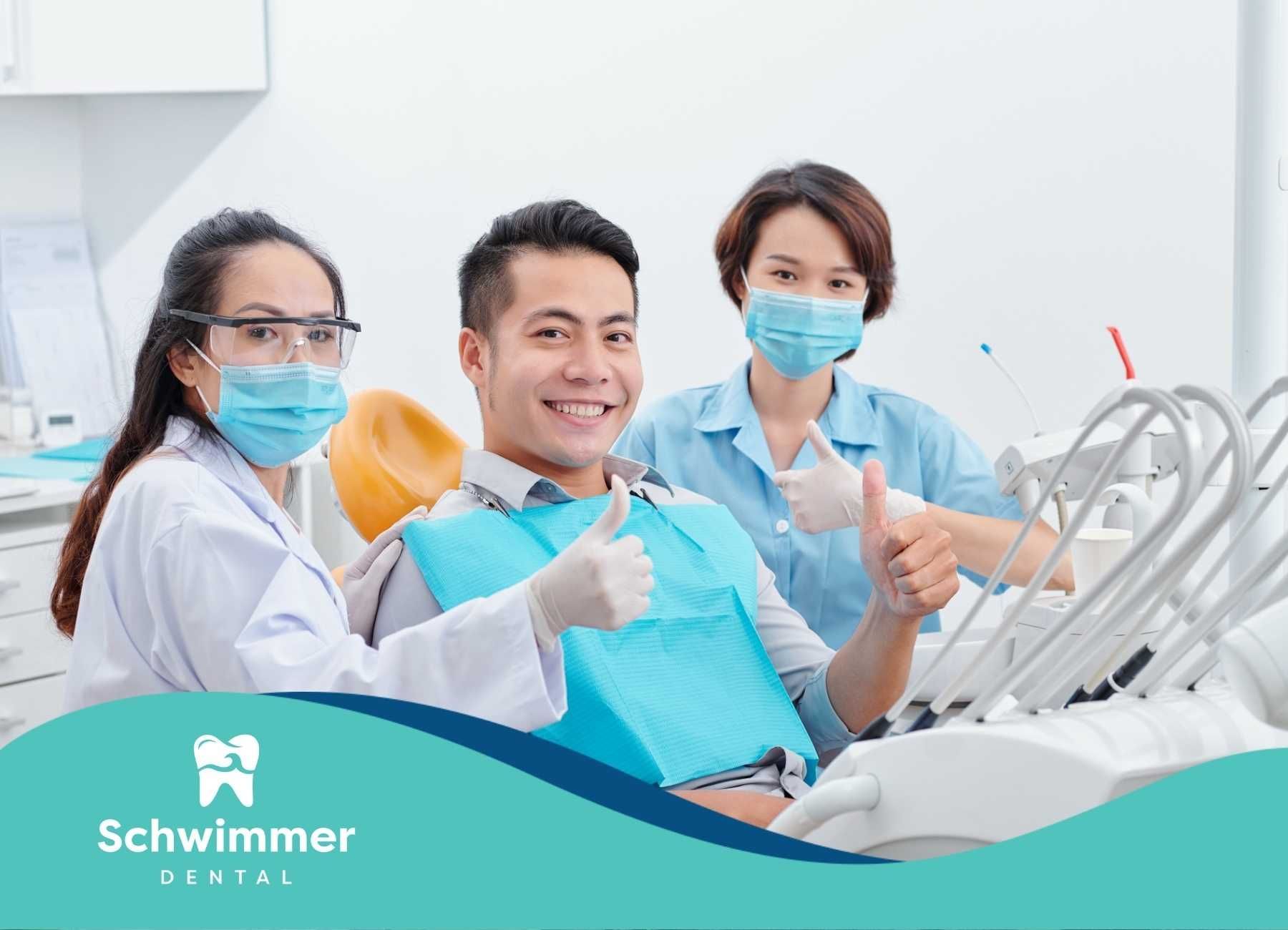 Smiling patient in dentist chair with two dental staff giving thumbs up.