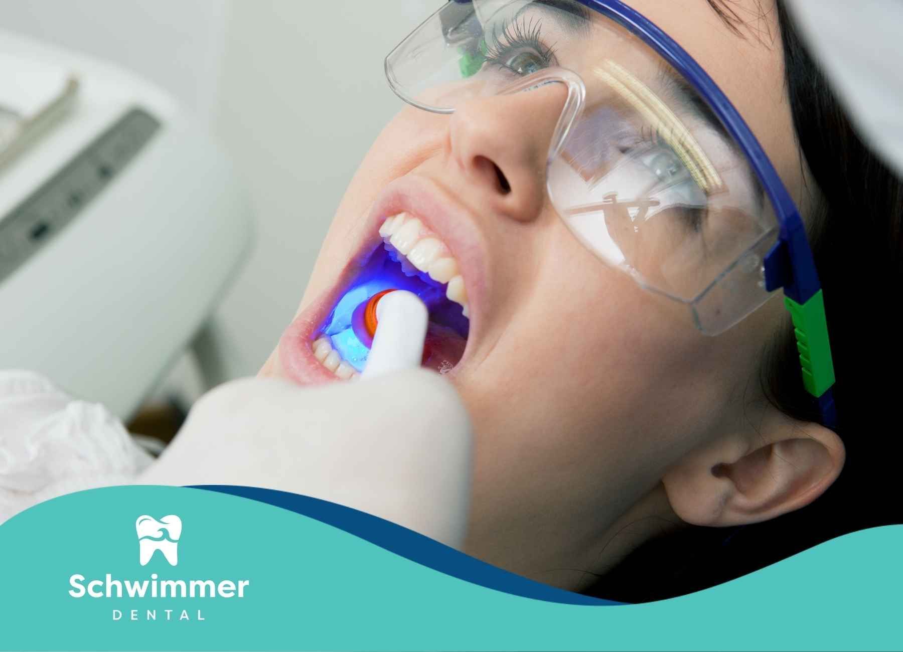 A dentist using a curing light on a patient's mouth. Patient wears safety glasses and has mouth open. Blue light shines.