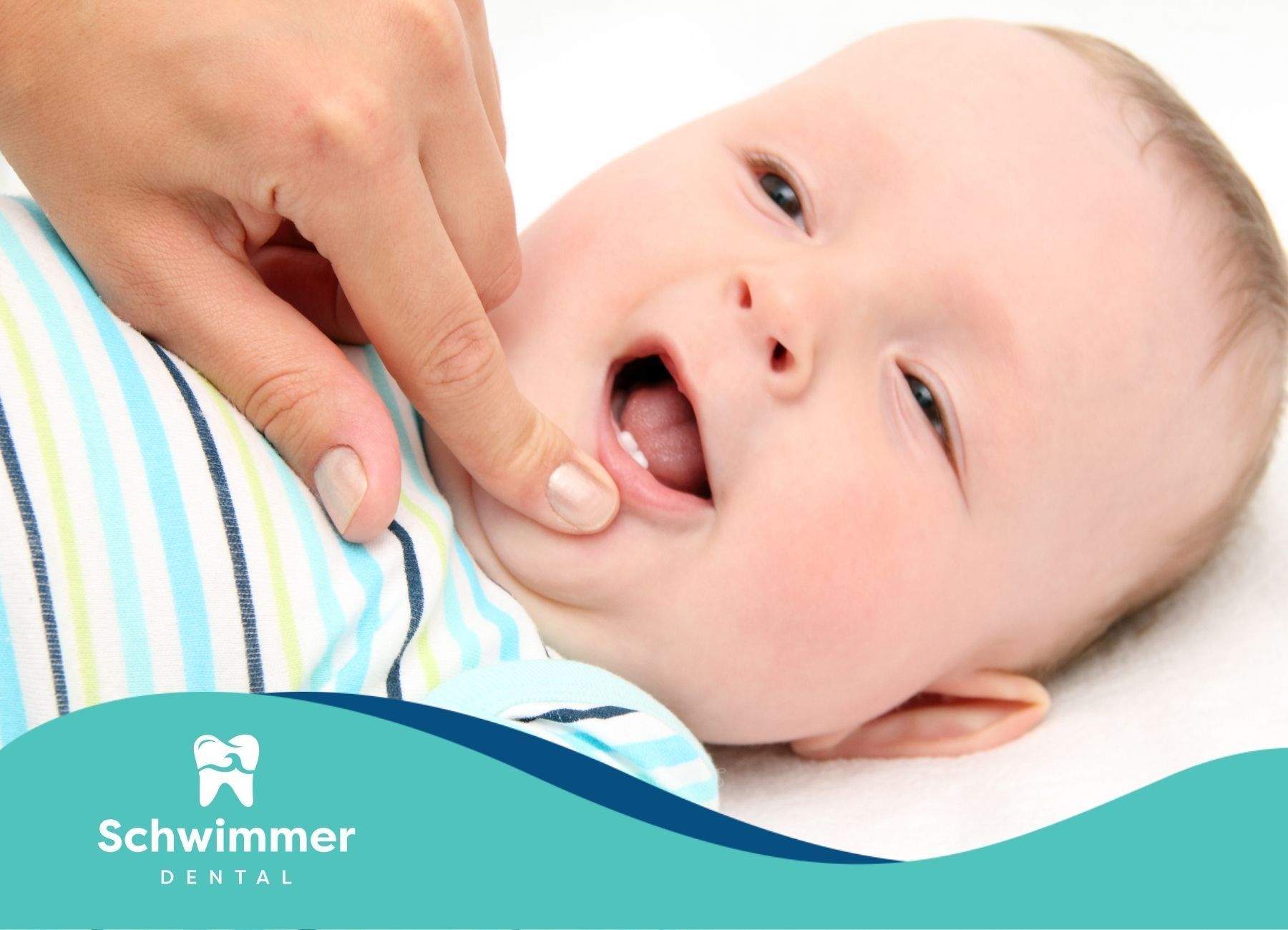 Baby's mouth open, finger pointing at emerged teeth, on a white surface, wearing striped shirt.