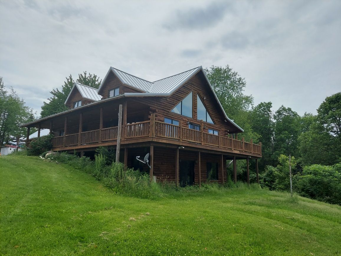 Log cabin home with wrap-around porch, sitting on a grassy hill, under a cloudy sky.