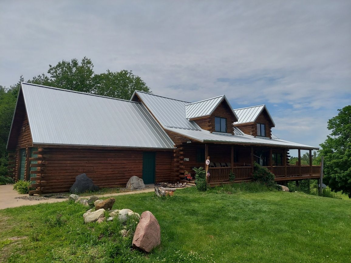 Log cabin with gray metal roof, porch, and green door on a grassy hill under a cloudy sky.