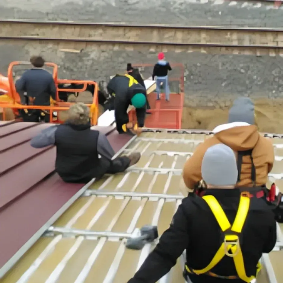 Construction workers installing a dark red roof near railroad tracks. Several wear harnesses and winter hats.