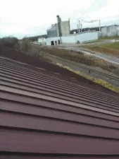 Red metal roof angled with a factory and train tracks in the background under a cloudy sky.