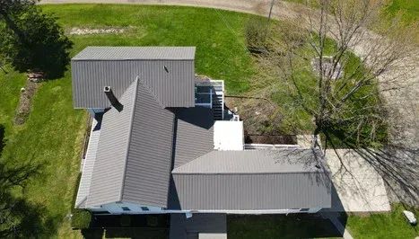 Overhead view of a house with a gray metal roof, chimney, and surrounding green lawn and driveway.