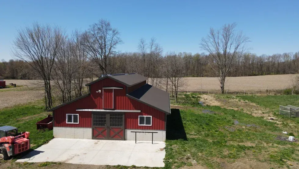 Red barn with brown roof on a concrete pad, set on a grassy hill in front of a treeline.