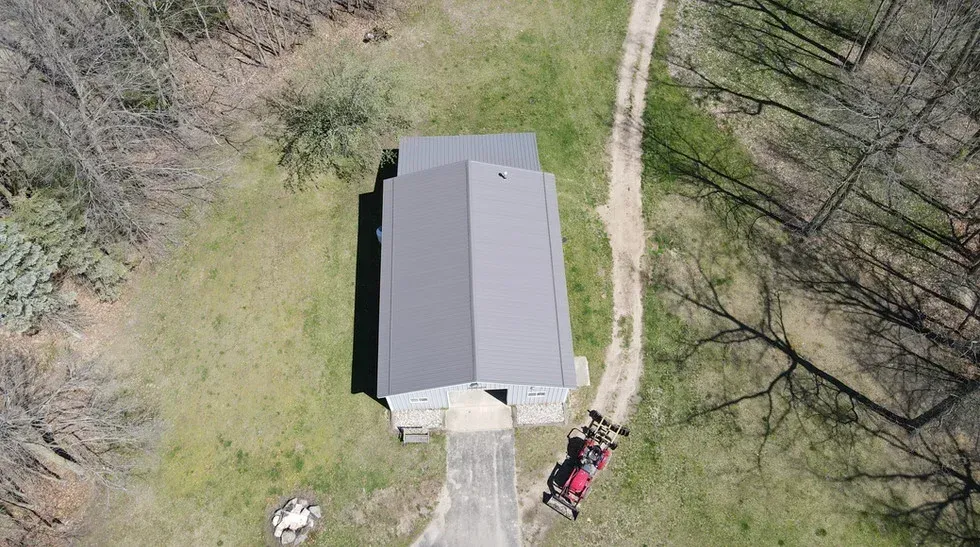 An aerial view of a gray-roofed house with a red vehicle parked on a concrete driveway, next to a dirt path.