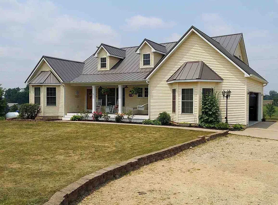 A light yellow house with a gray roof, porch, and a gravel driveway under a sunny sky.