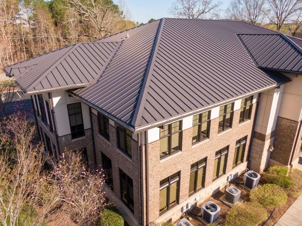 Multi-story brick building with gray metal roof; windows, air conditioning units, and landscaping.