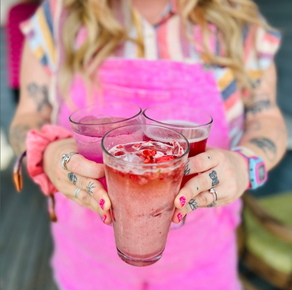 A woman in overalls is holding three glasses of pink liquid