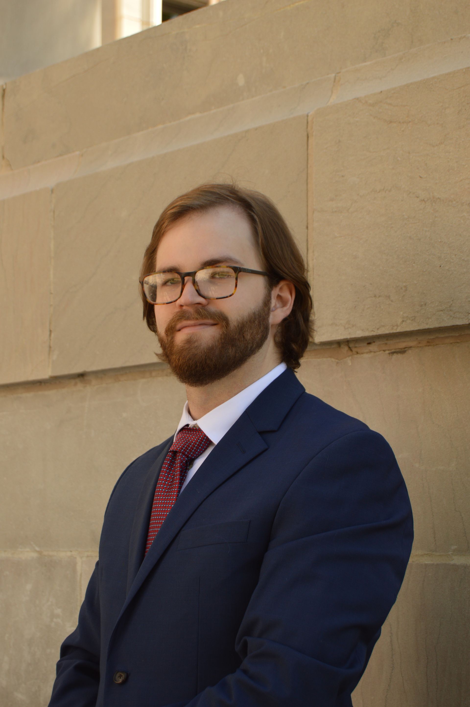 Man in a blue suit, glasses, and a red tie, leaning against a tan stone wall.