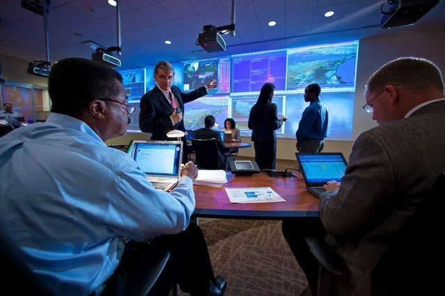 A group of people are sitting at a table with laptops in front of a large screen.