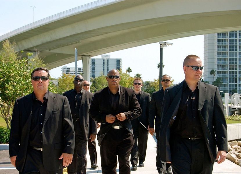 A group of men in suits and sunglasses are walking under a bridge.