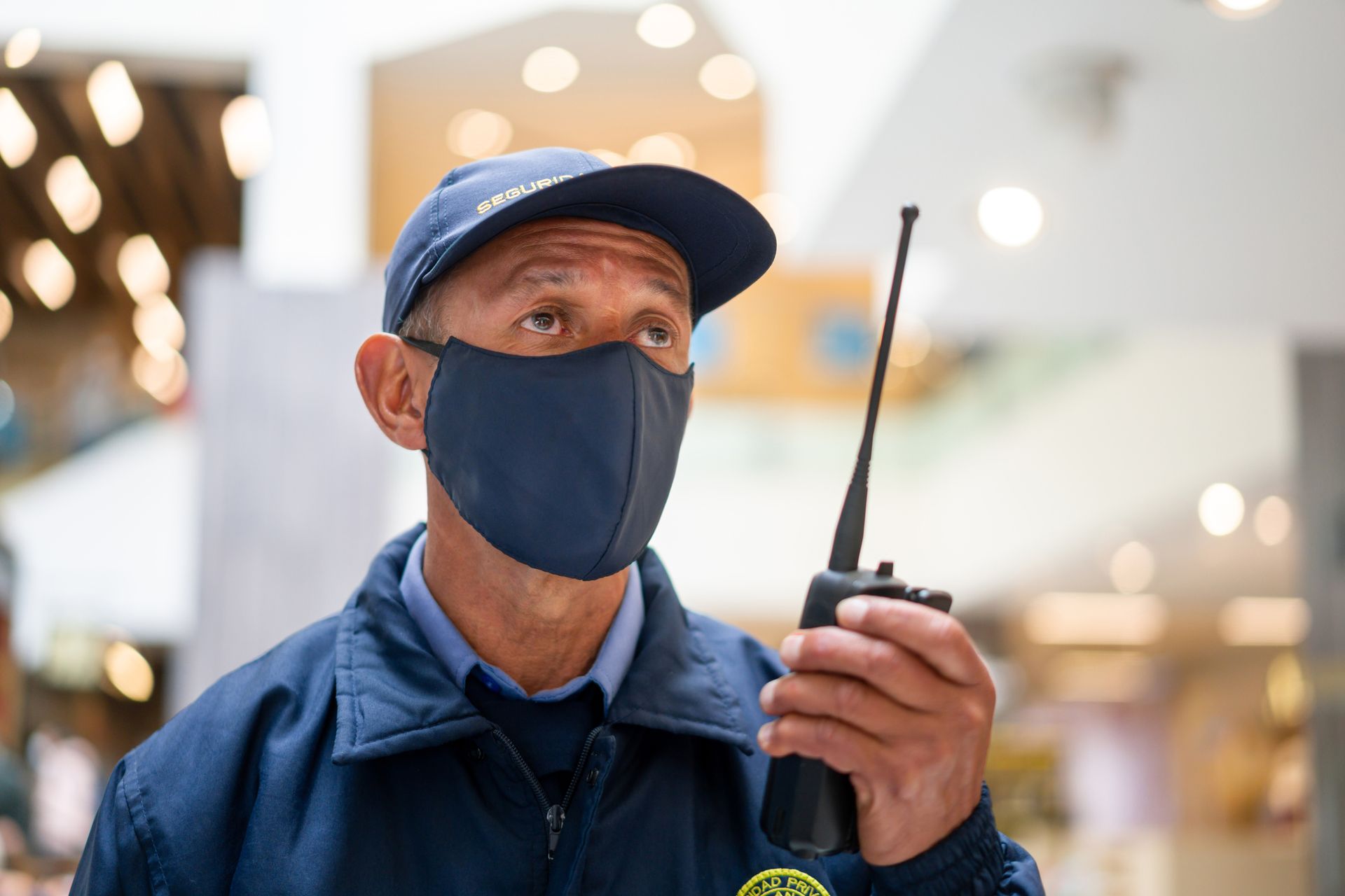 A security guard wearing a mask and a hat is holding a walkie talkie.