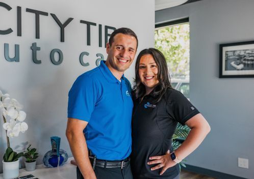 Blue Polo Shirt and a Woman in a Black Polo Shirt, Both Smiling, Stand Side-By-Side | Crown City Tire Auto Care