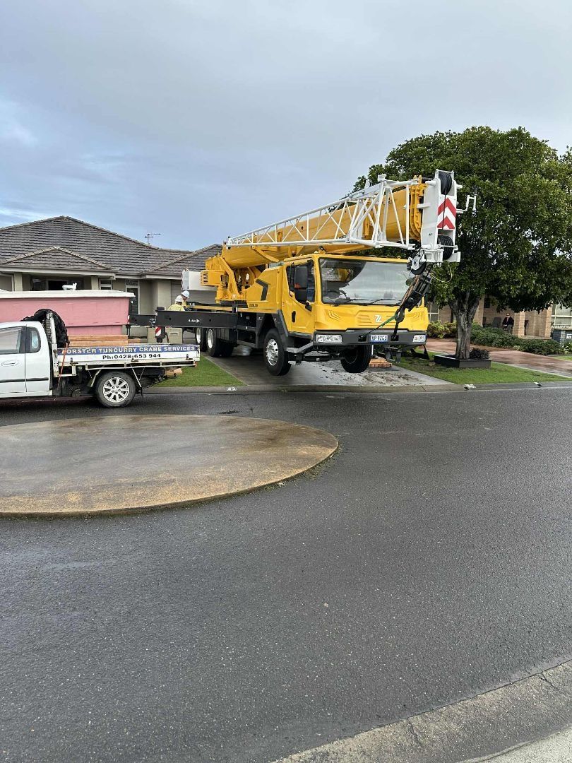 Yellow crane truck parked on a wet street next to a white pickup truck. Homes in background — Crane Rentals in Taree, NSW