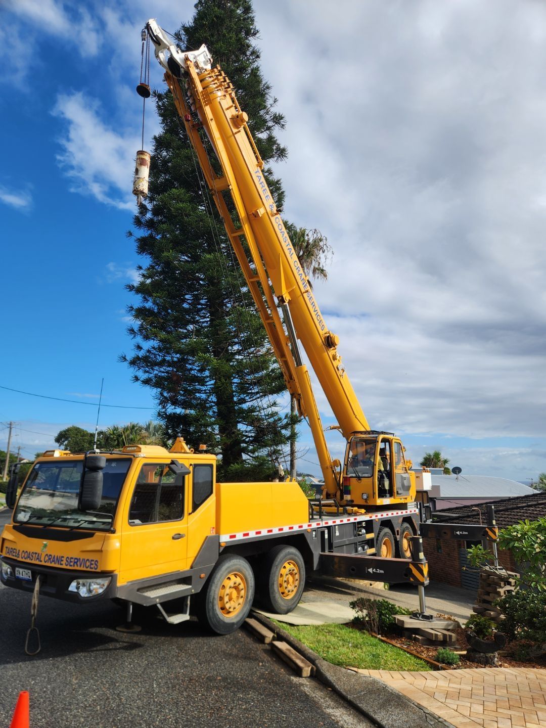 Yellow crane truck with extended boom lifting a load near a residential area — Crane Rentals in Taree, NSW