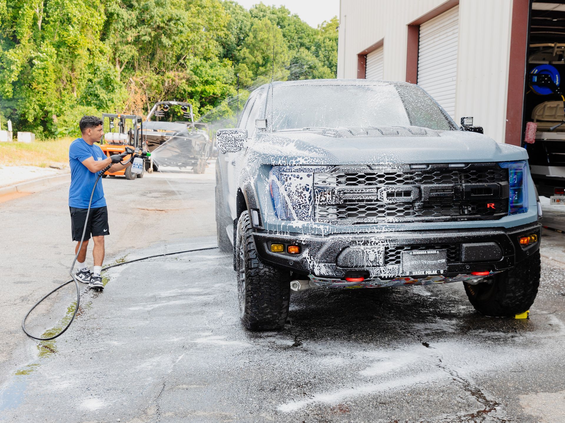 Man washing a grey truck with foam using a pressure washer outside a building.