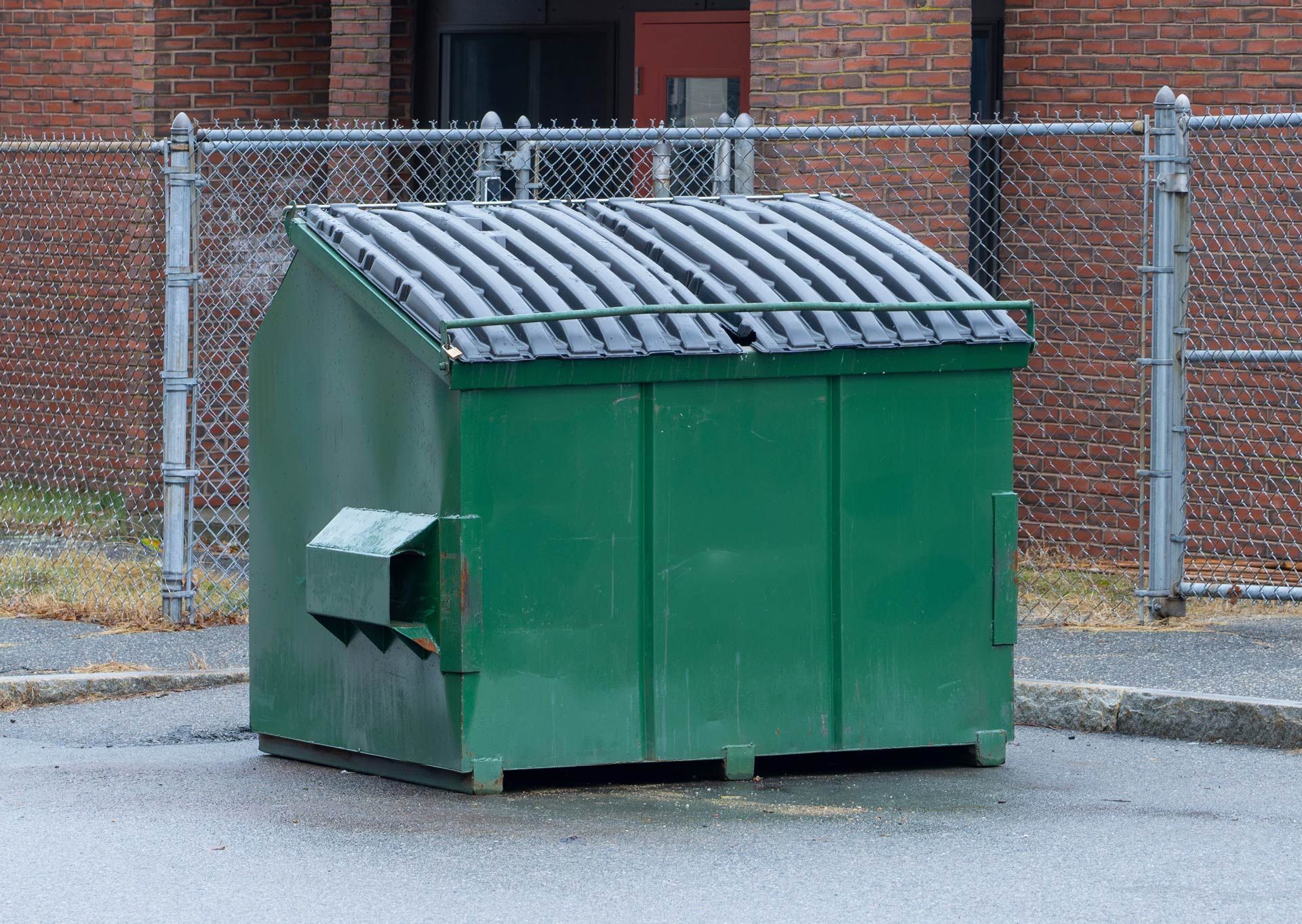 Green dumpster in front of a brick building behind a chain-link fence on a wet, paved surface.