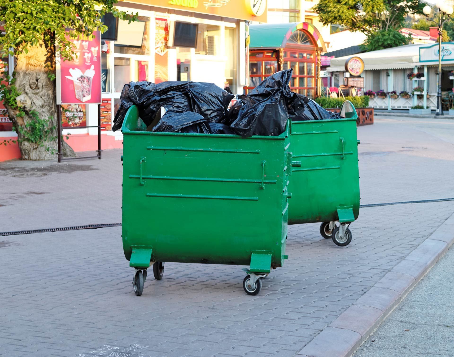 Two overflowing green dumpsters on a paved walkway, filled with black trash bags, in an outdoor setting.