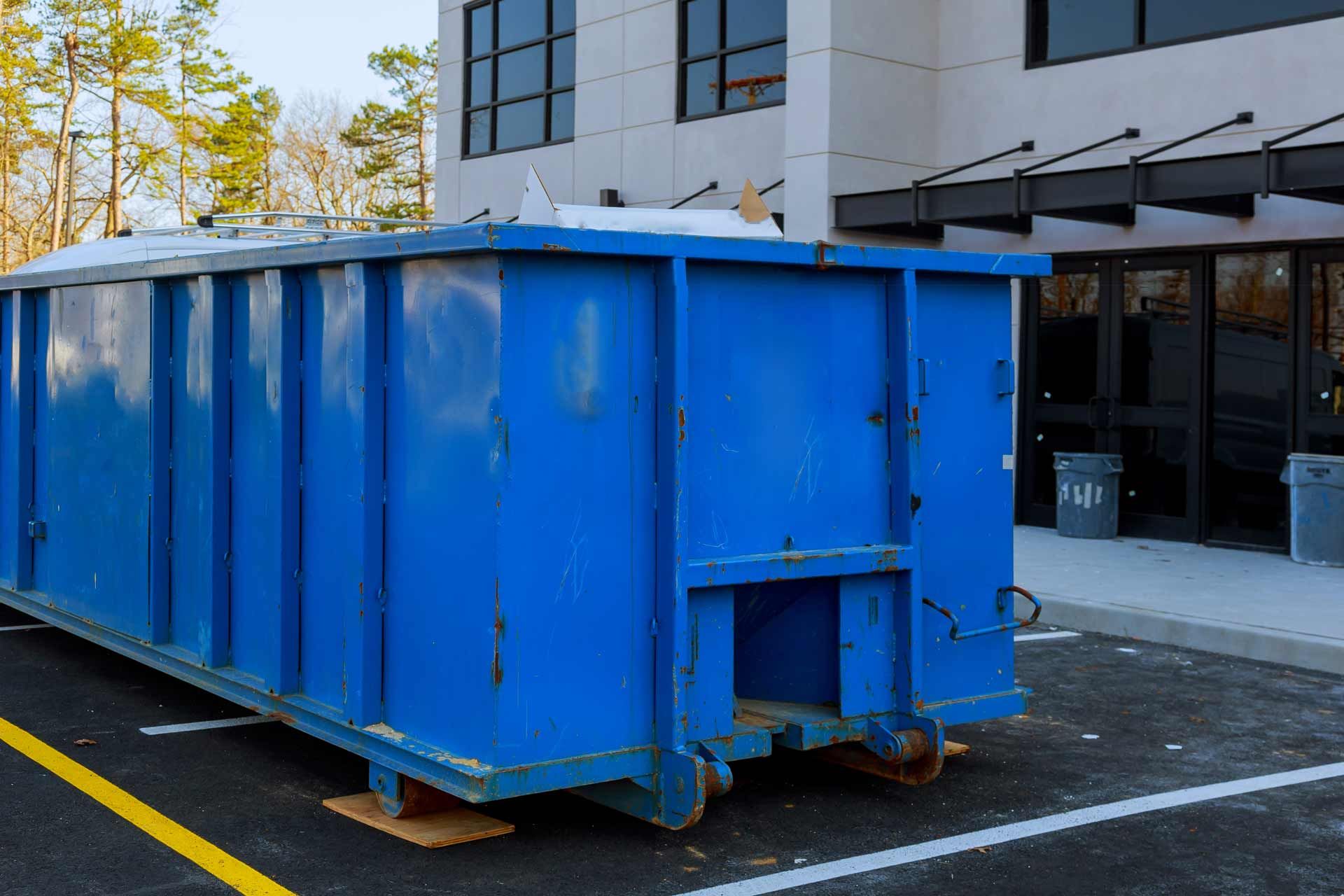 Blue dumpster sits outside a modern building with dark-framed windows.