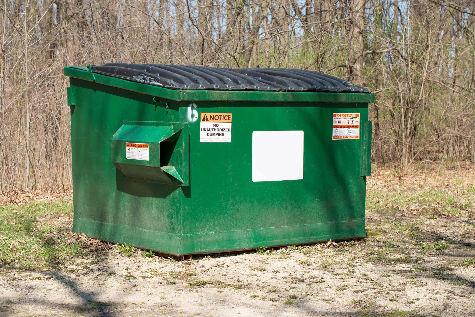 Green dumpster in a wooded area with a black lid and various warning stickers.