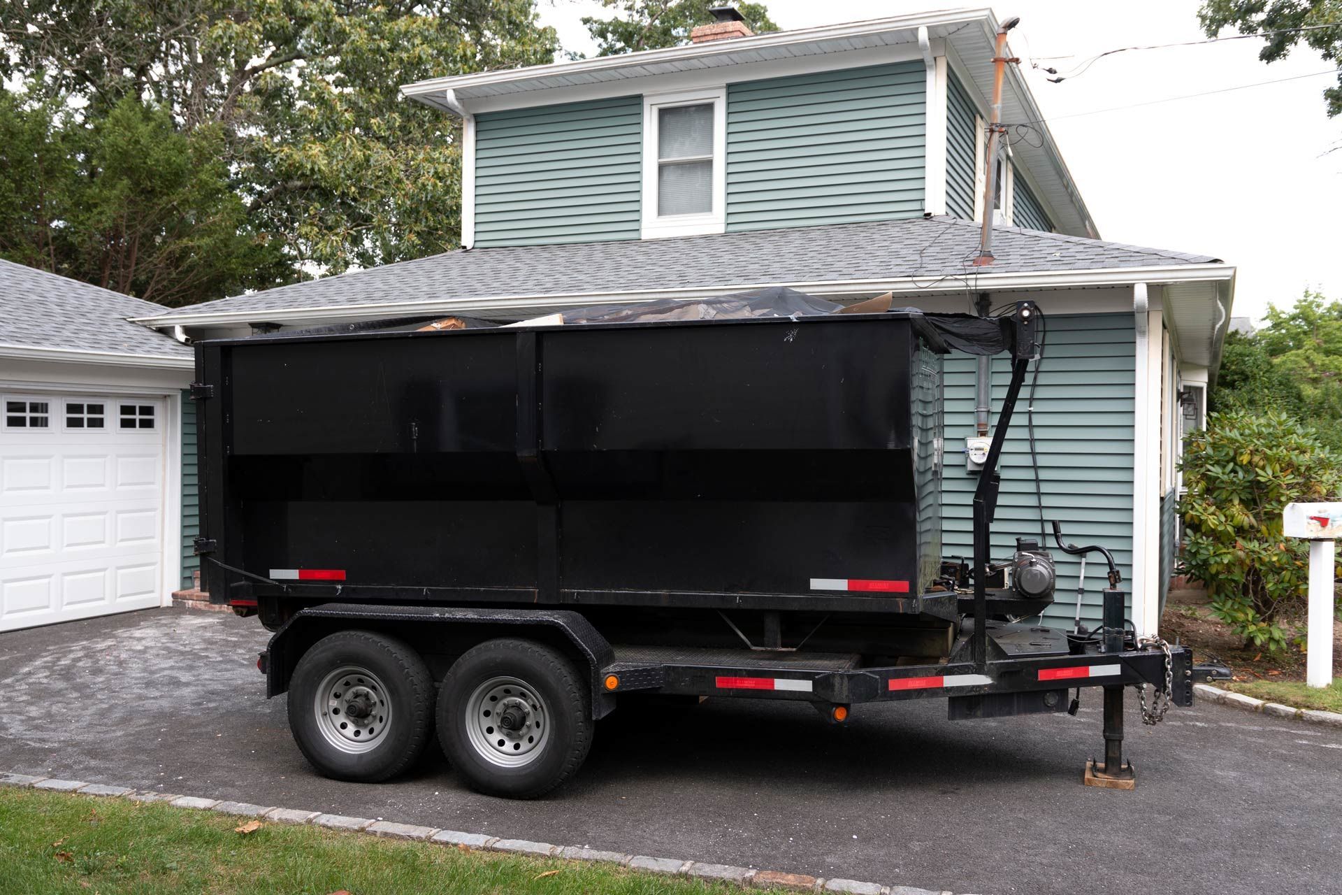 Black dumpster trailer parked in front of a blue house with a white garage.