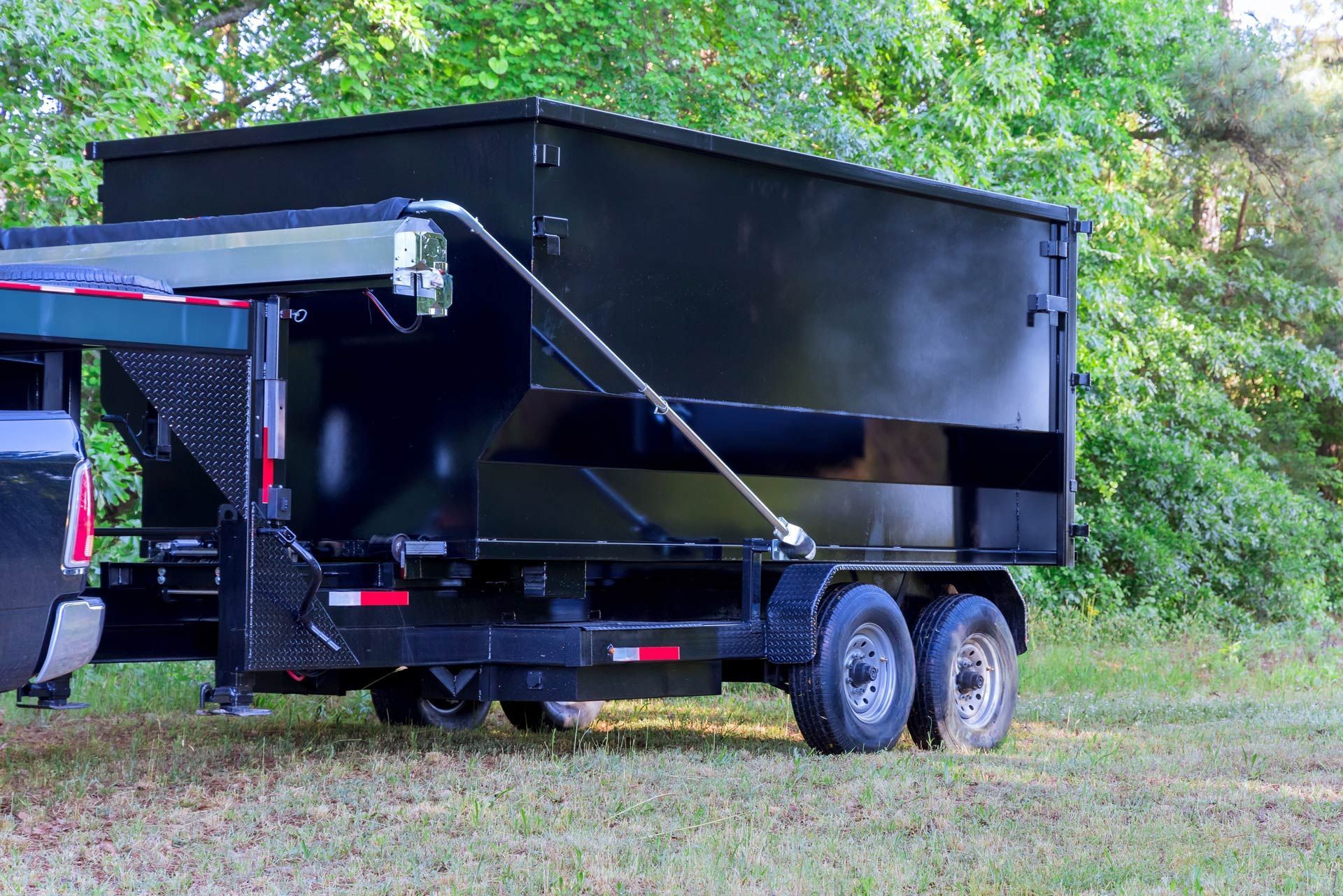 Black dump trailer hitched to a truck, in a grassy area with trees.