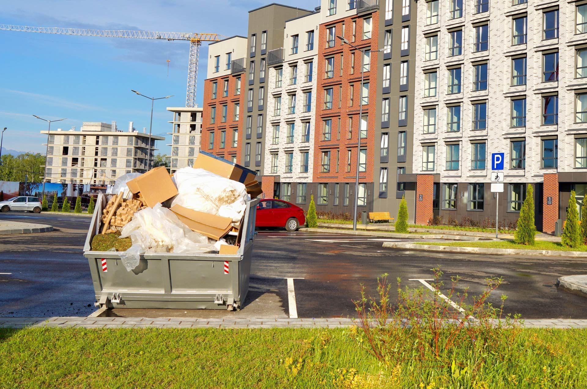 Full dumpster overflowing with cardboard and debris in a residential parking area, near buildings and construction.