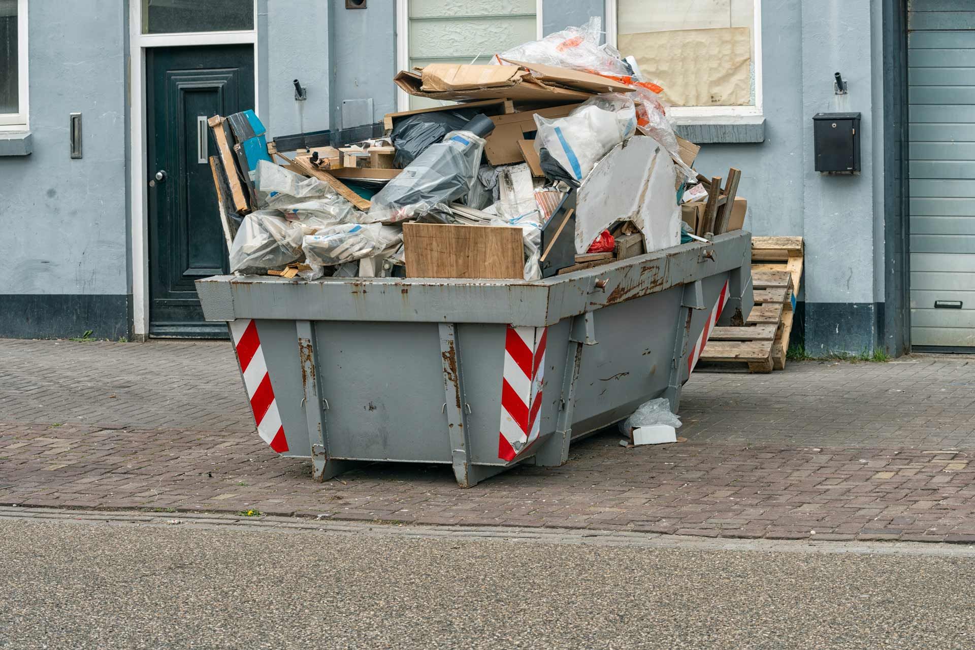 Gray dumpster overflowing with trash and debris on a street, against a blue building.