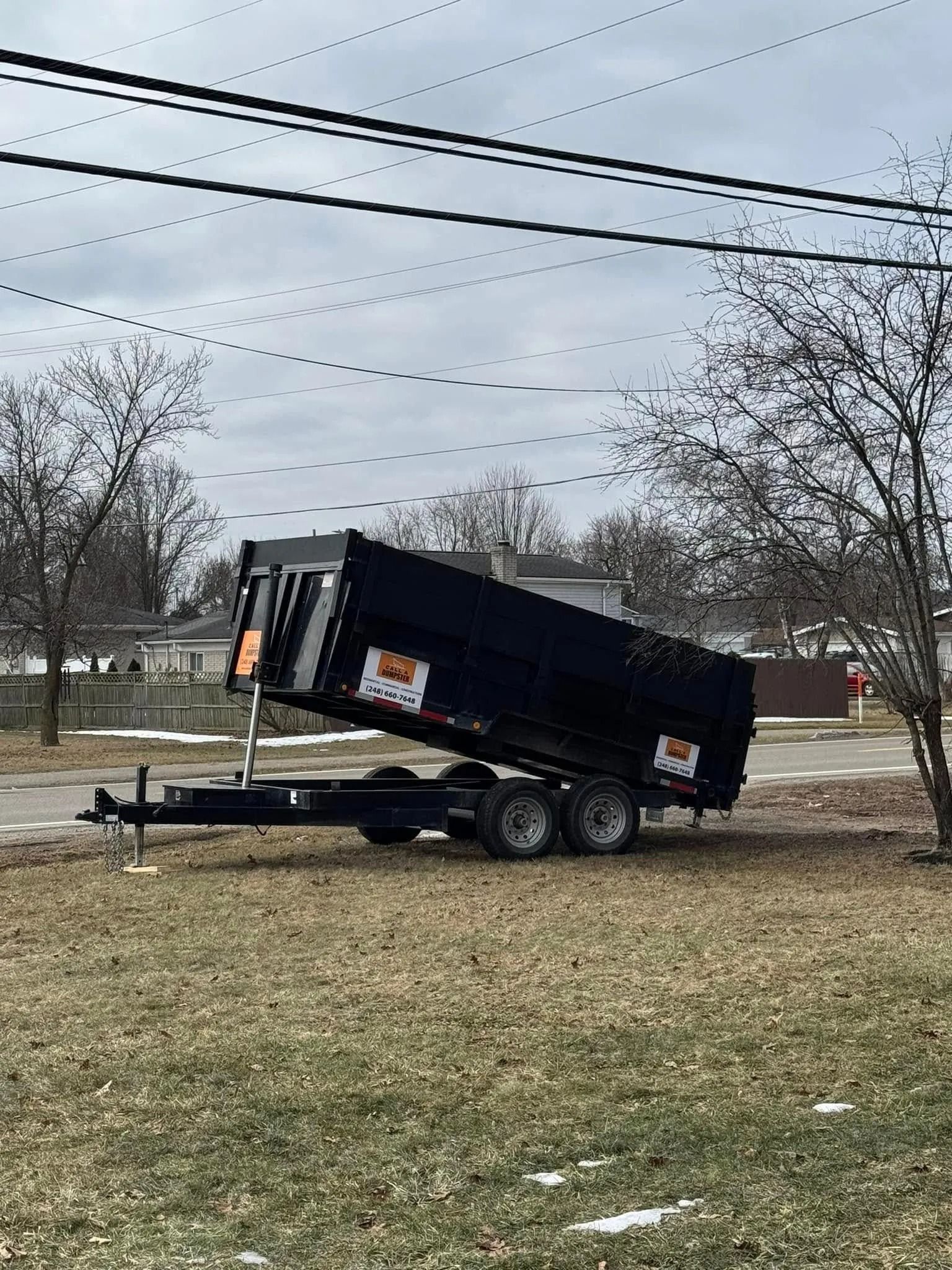 Black dumpster trailer hitched to a truck, in a grassy area with trees.