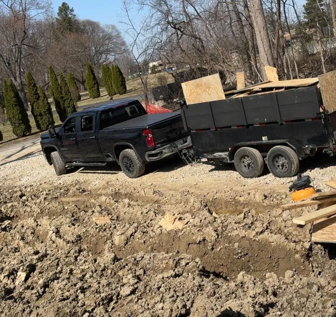 Black truck towing a trailer loaded with dark wood on a muddy construction site.