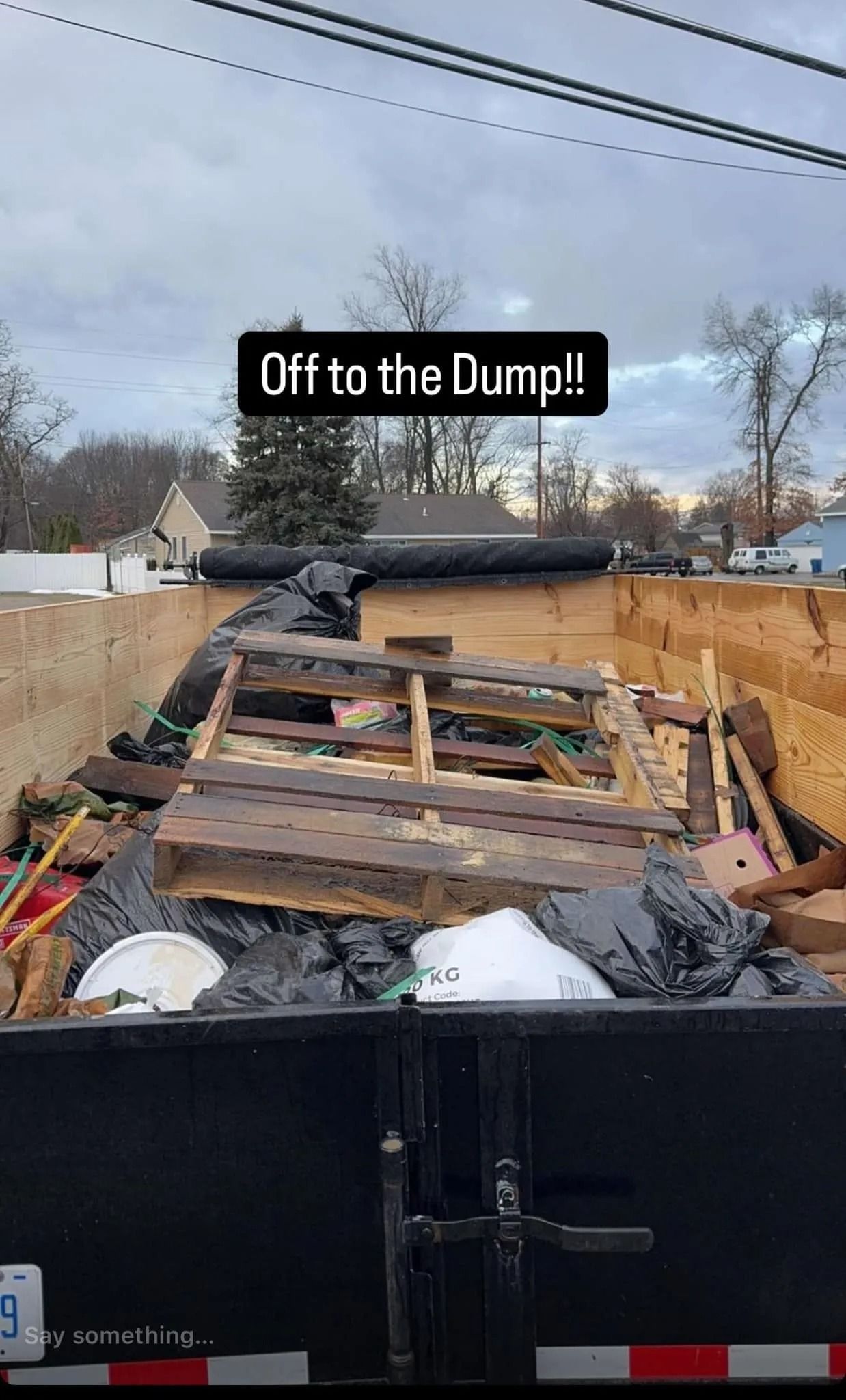 A dumpster overflowing with trash, including wood pallets, against a cloudy sky.
